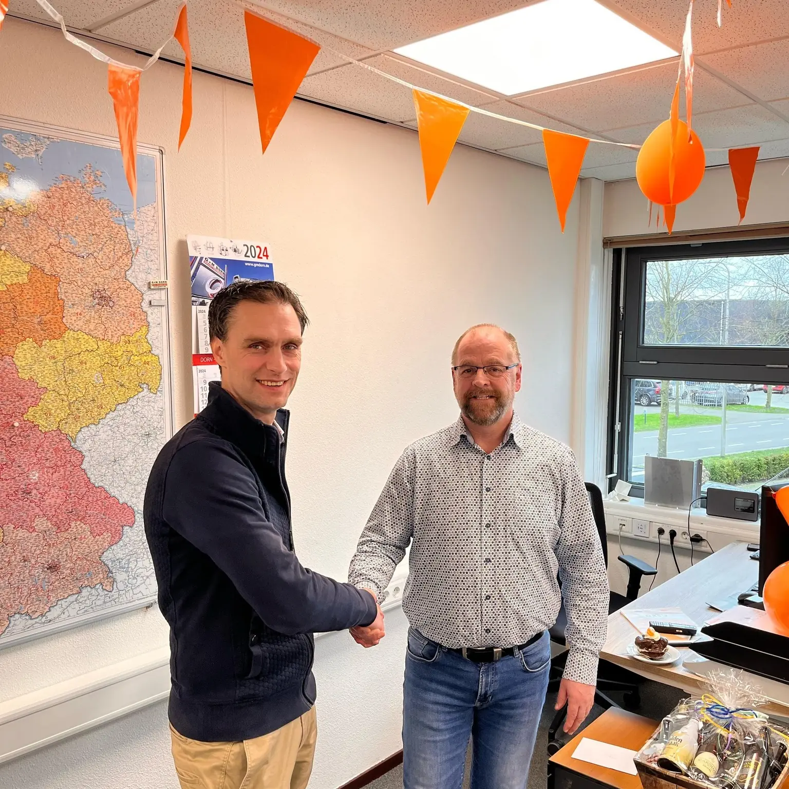 Rainer schudt de hand van een collega in een kantoor met oranje vlaggetjes